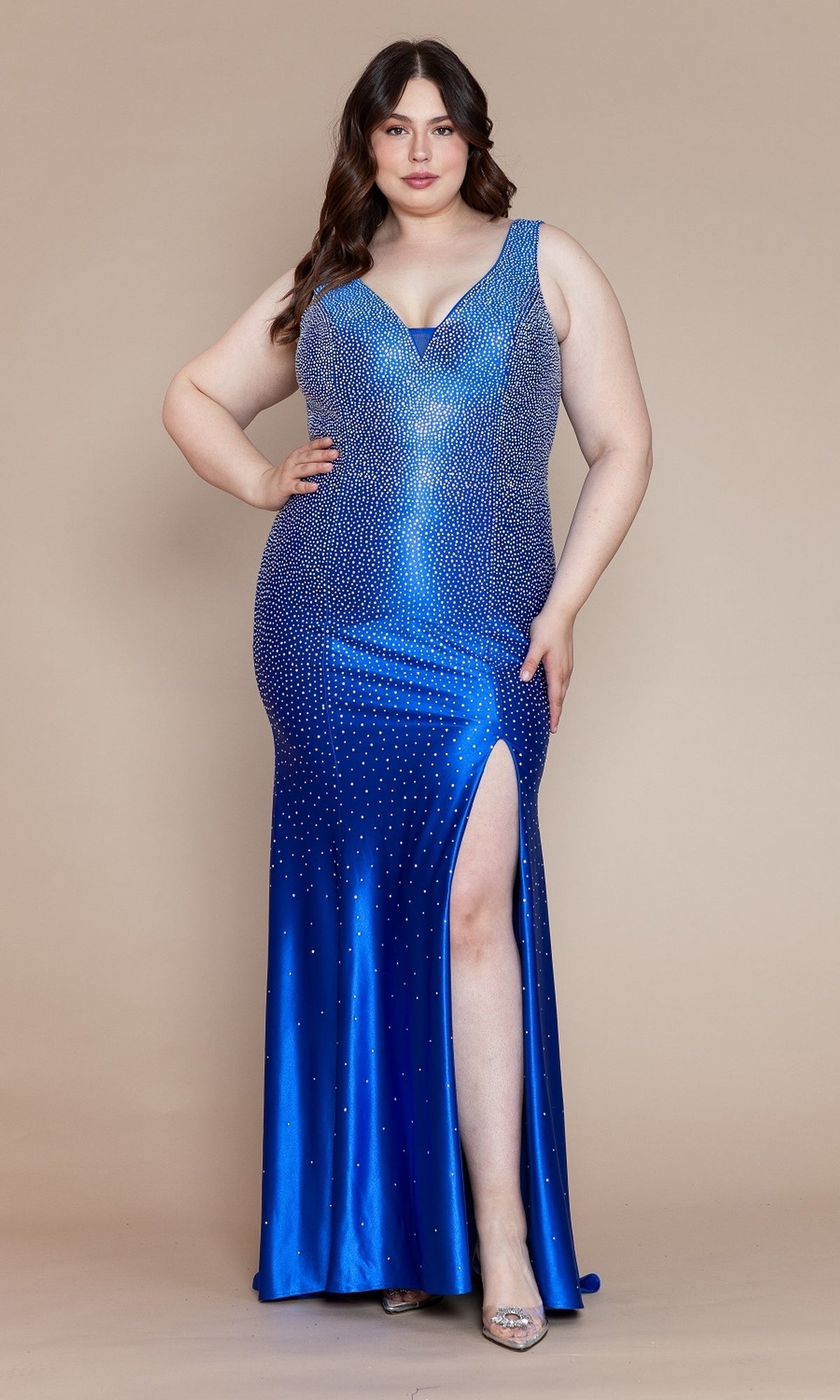 A woman wearing the Spandex Long Beaded Plus-Size Prom Dress W1130—a sleeveless, blue gown with a thigh-high slit and rhinestone embellishments—stands against a beige background.